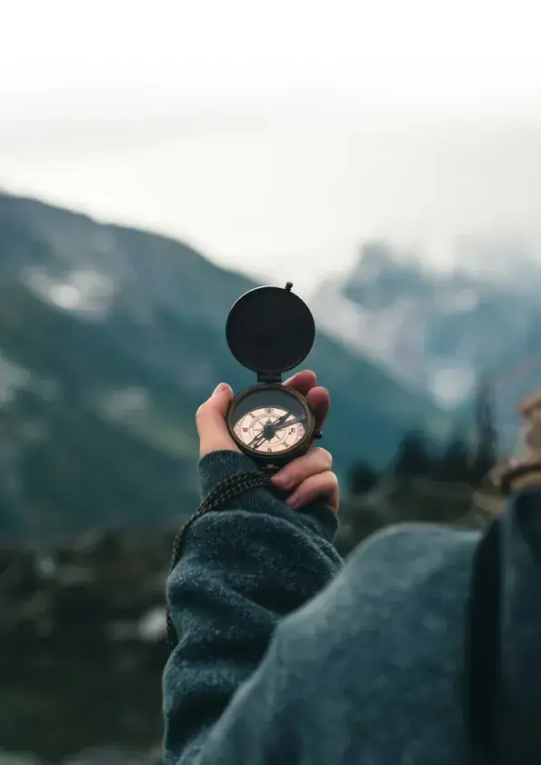 A hand holds an open compass against a mountain landscape.